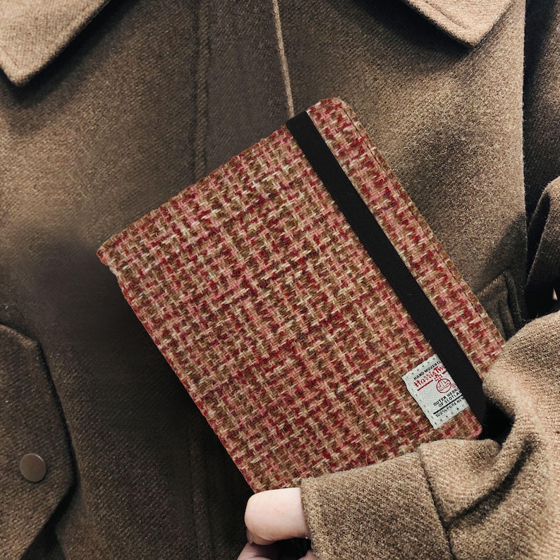 May include: A rectangular notebook with a red, brown, and white tweed cover. A black elastic band secures the notebook. A small label is visible on the bottom right corner. The notebook is held against a brown coat.