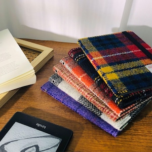 May include: A stack of colorful plaid fabric book covers in various colors, including red, blue, yellow, orange, and purple. A Kindle e-reader is in the foreground, and a book and frame are in the background.