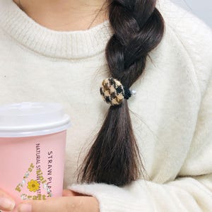 May include: A brown, black, and tan plaid fabric-covered hair tie secures a dark brown braid. A small white pearl accentuates the tie. The person is wearing a white sweater and holding a pink coffee cup with text.