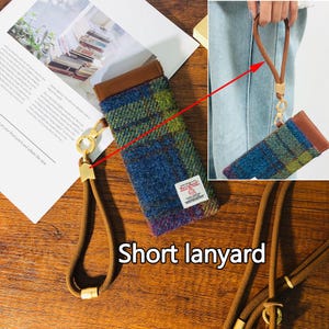 May include: A blue, green, and brown plaid phone case with a brown leather top and a short lanyard. The lanyard is a tan color with gold-colored metal accents. The case has a small white tag with red text.