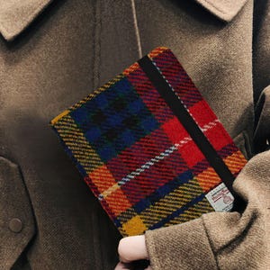Könnte beinhalten: Ein bunt kariertes Notizbuch mit einem schwarzen Gummibandverschluss. Das Notizbuch hat ein lebendiges Muster aus Rot, Blau, Gelb und Grün. Ein kleines Etikett mit Text ist sichtbar.