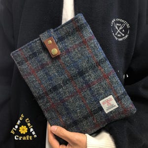 Peut inclure: Une pochette pour tablette en tissu gris et bleu à carreaux avec un rabat en cuir marron et un bouton en métal doré. La pochette porte une petite étiquette avec le texte "Harris Tweed".