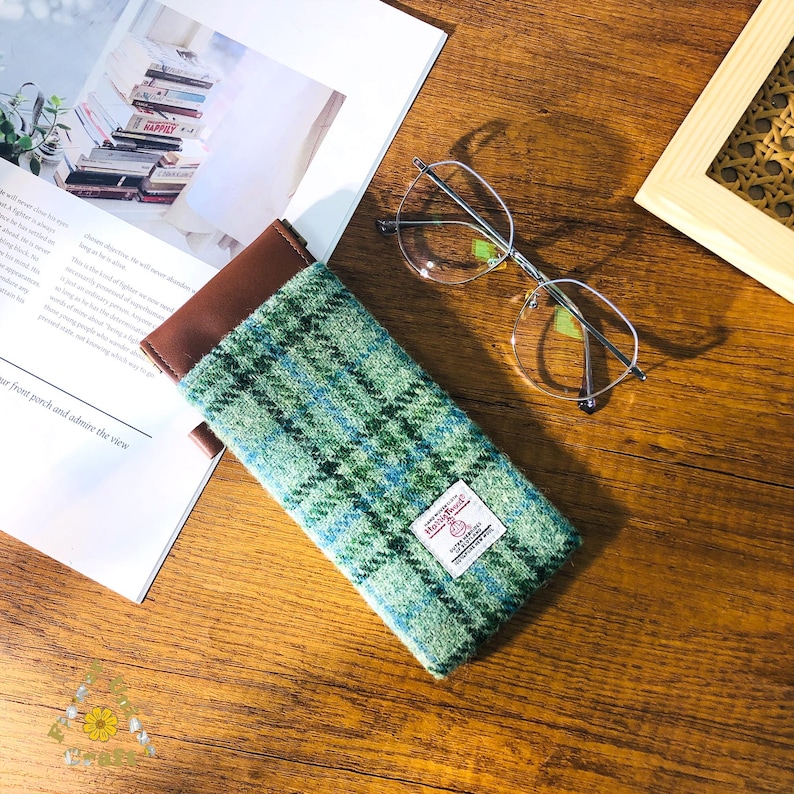 May include: A green and blue plaid eyeglasses case with a brown leather top. The case is rectangular and has a small white tag with text. A pair of silver-rimmed glasses rests nearby on a wooden surface.