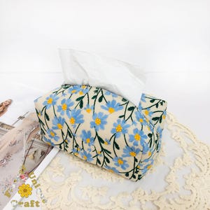May include: A white tissue box cover with a floral pattern. The cover is made of fabric and has a light beige background with blue and yellow flowers.