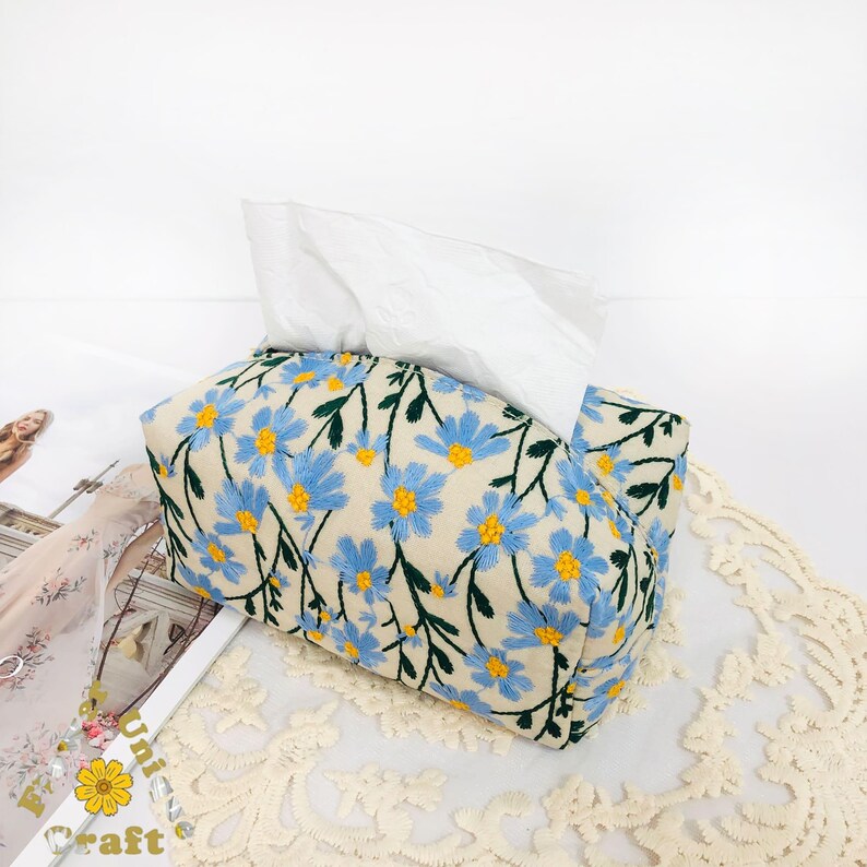 May include: A white tissue box cover with a floral pattern. The cover is made of fabric and has a light beige background with blue and yellow flowers.