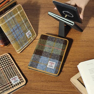 May include: A collection of rectangular cases featuring Harris Tweed wool fabric in various plaid patterns, including olive, blue, and brown tones. Each case has a small white label with red text. One case is propped open on a black stand.