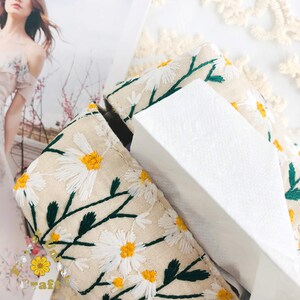 May include: A white tissue box cover with a floral design. The cover is made of beige fabric with embroidered white daisies and green stems.