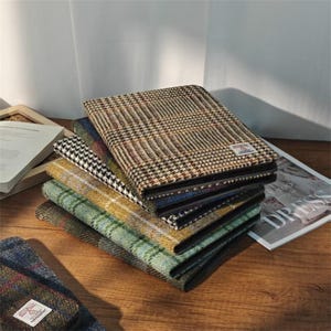 Puede incluir: Una pila de cuadernos con cubiertas de tweed estampadas en varios colores, incluyendo marrón, negro, verde y amarillo. Los cuadernos están colocados sobre una superficie de madera, con una revista y un libro en el fondo.