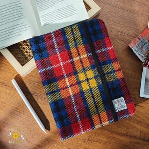 Puede incluir: Funda rectangular para tableta a cuadros multicolores con una banda elástica negra. La funda presenta un patrón de rojo, azul, amarillo y blanco. También se ve un lápiz óptico blanco y un libro abierto.