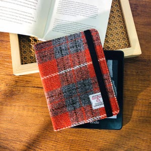 Puede incluir: Una funda Kindle a cuadros rojos, grises y blancos con una banda elástica negra. La funda está hecha de una tela texturizada y está sobre un dispositivo Kindle negro. Un libro y un marco de madera están en el fondo.