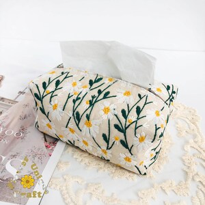 May include: A beige tissue box cover with an embroidered design of white daisies and green leaves.