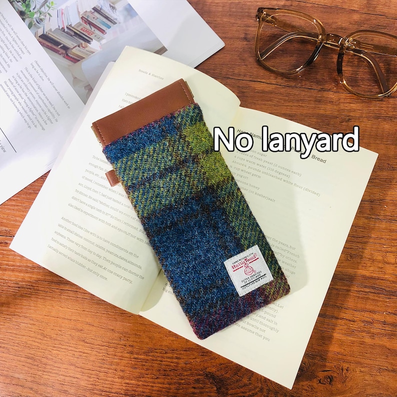 May include: A brown leather and tweed glasses case with a red and white label that says "Harris Tweed" and "Hand Woven." The case is lying on a white page with the text "No lanyard" written on it.