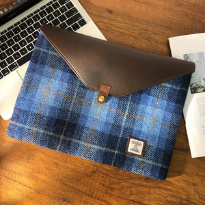 Op de afbeelding: Blauwe en bruine geruite laptophuls met een leren klep en een messing sluiting. De hoes heeft een klein label met de tekst "Harris Tweed".