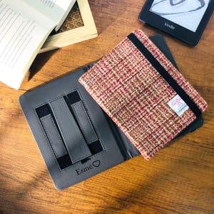 May include: A gray tablet case with a pink, red, and brown tweed cover. The case has a black elastic band and a small label. The word "Esm&eacute;" and a heart are printed on the case. A Kindle e-reader and a book are also visible.