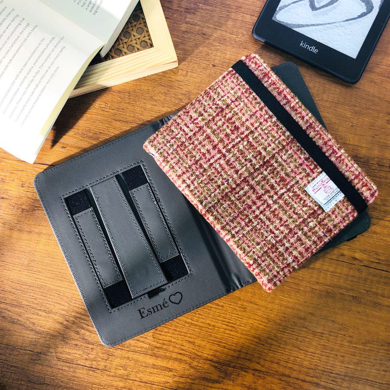 May include: A gray tablet case with a pink, red, and brown tweed cover. The case has a black elastic band and a small label. The word "Esm&eacute;" and a heart are printed on the case. A Kindle e-reader and a book are also visible.