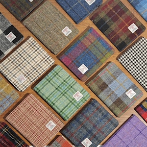 May include: An assortment of rectangular tweed wallets in various colors and patterns, including plaid, herringbone, and houndstooth. Each wallet features a small white label with text. The wallets are arranged on a wooden surface.