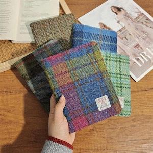 Op de afbeelding: Een hand die een veelkleurige notitieboekhoes met ruitpatroon vasthoudt. De hoes heeft een mix van blauw, paars, groen en oranje. Andere notitieboekhoezen met verschillende tweedpatronen zijn zichtbaar op de achtergrond. Een tijdschrift is ook zichtbaar.