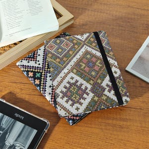 May include: A patterned journal with a black elastic band closure. The cover features a geometric design in shades of purple, green, and cream. The journal is lying on a wooden surface next to a Kindle and an open book.