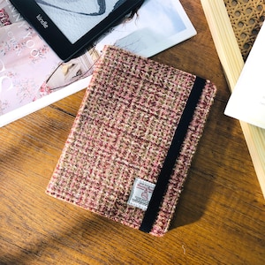 May include: A rectangular book cover made of a textured, woven fabric in shades of pink, brown, and green. A black elastic band secures the cover. A small label is sewn on the cover. The cover is on a wooden surface.
