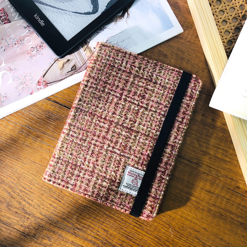 May include: A rectangular book cover made of a textured, woven fabric in shades of pink, brown, and green. A black elastic band secures the cover. A small label is sewn on the cover. The cover is on a wooden surface.