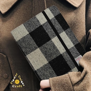 Puede incluir: Un cuaderno con un patrón de cuadros en blanco, negro y gris. Tiene un cierre de banda elástica negra y una cubierta de tela. El cuaderno lo sostiene una persona que lleva una chaqueta marrón. La cubierta del cuaderno es de tela texturizada.