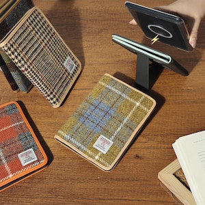May include: A collection of rectangular phone cases featuring Harris Tweed wool fabric in various plaid patterns, including olive green, blue, and orange. Each case has a small white label and a black stand, with one case shown charging wirelessly.
