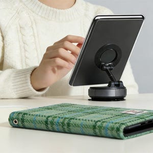 以下が含まれることがあります： 黒いタブレットが吸盤ベースのスタンドに取り付けられています。緑のチェック柄のタブレットケースが前景にあります。ケースはテクスチャード加工された生地と小さなラベルが付いています。背景には白いセーターを着た人が写っています。
