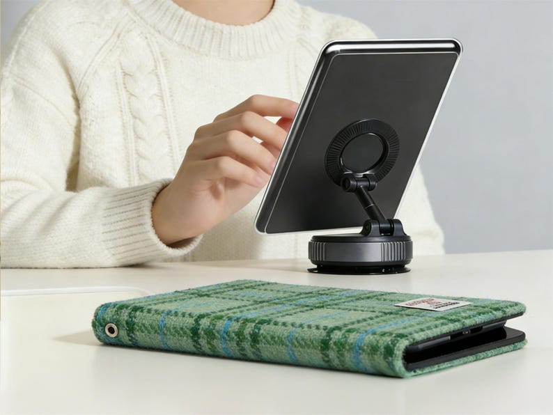以下が含まれることがあります： 黒いタブレットが吸盤ベースのスタンドに取り付けられています。緑のチェック柄のタブレットケースが前景にあります。ケースはテクスチャード加工された生地と小さなラベルが付いています。背景には白いセーターを着た人が写っています。
