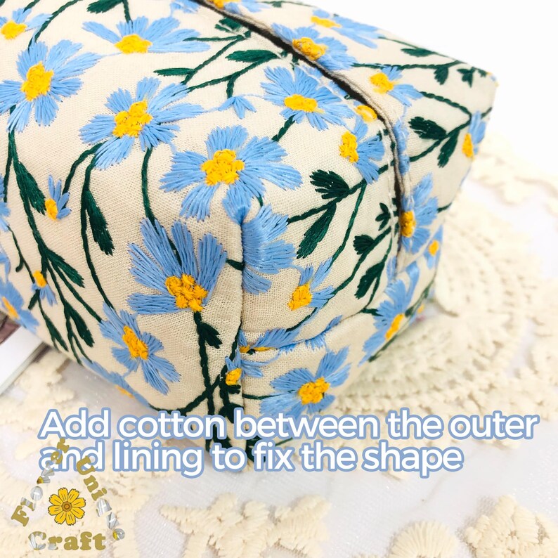 May include: A close-up of a beige fabric pouch with blue and yellow embroidered flowers. The pouch is sewn with a seam along the top edge. The text "Add cotton between the outer and lining to fix the shape" is visible at the bottom of the image.