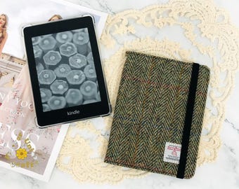 May include: A green, brown, and red tweed Kindle e-reader case with a black elastic closure. The case has a small label with the text "Harris Tweed" on it.