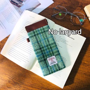 May include: A green and blue plaid glasses case with a brown leather top and a small label. The case is resting on an open book, with a pair of eyeglasses and a wooden block visible in the background.