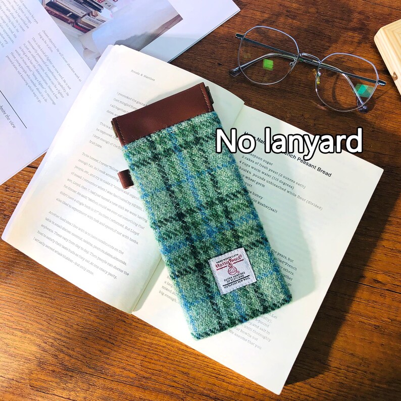 May include: A green and blue plaid glasses case with a brown leather top and a small label. The case is resting on an open book, with a pair of eyeglasses and a wooden block visible in the background.