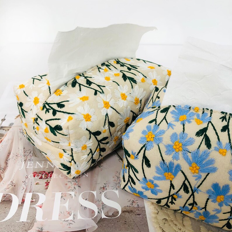May include: Two tissue box covers with floral embroidery. One cover features white daisies on a cream background, while the other has blue daisies on a cream background.
