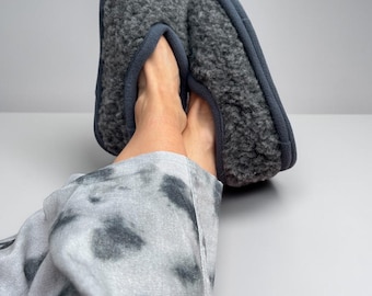 May include: A pair of dark grey slippers featuring a textured, curly fleece exterior and a smooth, solid dark grey trim along the opening. The slippers have a rounded toe design and a soft, plush appearance, providing a simple and functional look.