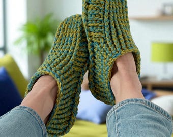 May include: A pair of handmade, knitted slippers in shades of olive green and teal blue. The slippers have a ribbed texture and are designed to cover the entire foot. The person wearing the slippers is also wearing blue jeans.