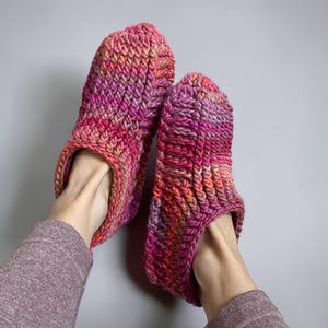 May include: A pair of handmade, chunky knit slippers in vibrant shades of pink, orange, and purple. The slippers have a textured, ribbed design and are shown on a gray background. These cozy slippers are perfect for indoor wear.