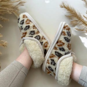 May include: A pair of brown and white patterned slippers with a white fleece lining. The slippers are being worn on a person's feet.