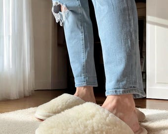 May include: A pair of cream-colored slippers featuring a plush, textured fleece upper and a smooth, rounded toe design. The slippers are worn with light blue distressed denim jeans that feature visible rips at the knees and frayed edges.