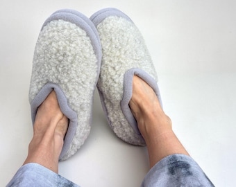 May include: A pair of slip-on slippers featuring a textured, off-white faux shearling upper with a soft lavender trim along the collar and opening. The slippers have a rounded toe design and a plush, fleece-like interior lining for comfort.