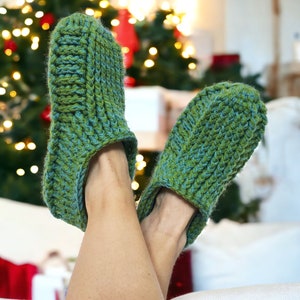 Op de afbeelding: Een paar groene gehaakte pantoffels met een textuurontwerp. De pantoffels worden gedragen door iemand met hun voeten op een witte bank.