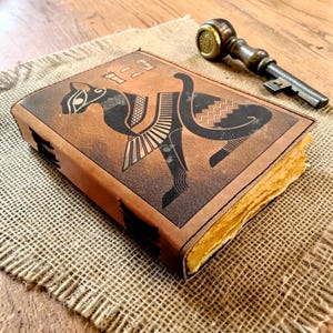 May include: A brown leather journal with an embossed black cat design and hieroglyphs. The journal has aged, yellowed pages and is accompanied by a vintage key with a brass handle. The journal is on a burlap surface.
