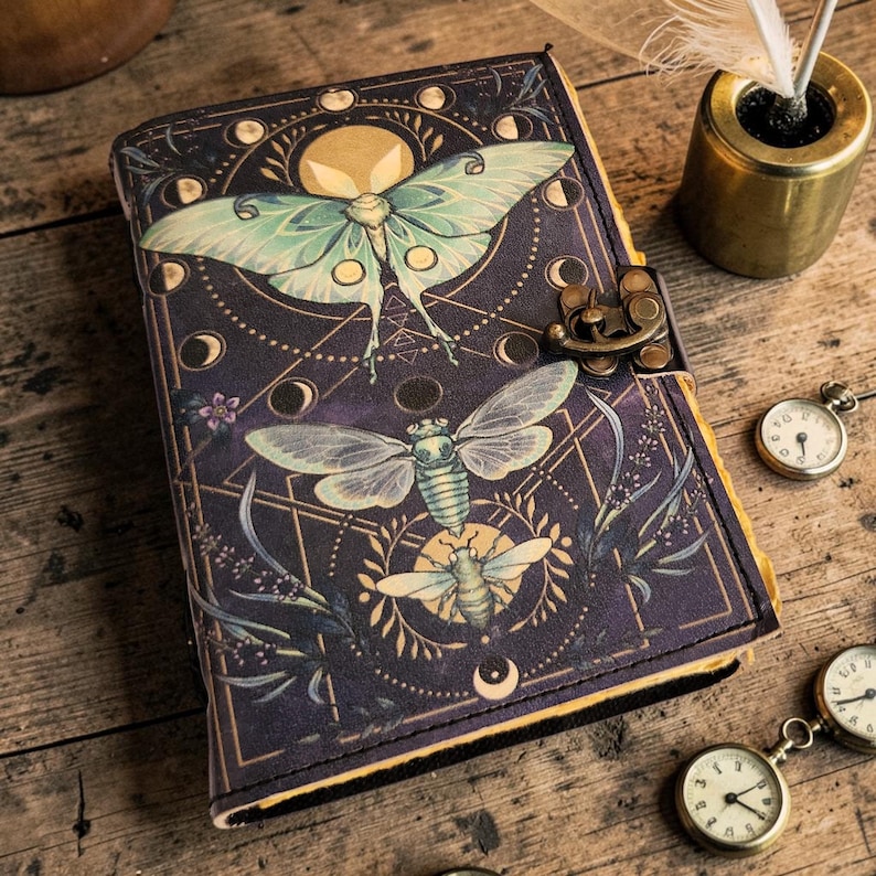 Puede incluir: Un diario azul oscuro con un dise&ntilde;o detallado de insectos y celestial. La portada presenta una gran polilla y otros insectos, con fases lunares y patrones geom&eacute;tricos. Un cierre de metal asegura el diario, y peque&ntilde;os relojes de bolsillo est&aacute;n dispersos.