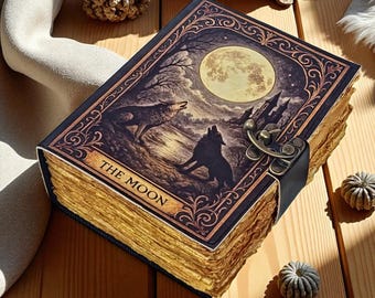 Puede incluir: Un diario encuadernado en cuero con una ilustración detallada de lobos aullando a la luna. La portada presenta las palabras "THE MOON" y se asegura con un cierre de metal y una correa de cuero. Las páginas tienen un borde dorado.