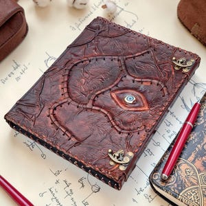 May include: A brown leather journal with a textured cover and stitched details. The cover features an eye design with a blue iris and is secured with a metal clasp. A red pen rests nearby, suggesting the journal is for writing.