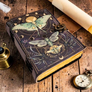 Puede incluir: Un diario de cuero morado oscuro con un diseño detallado de insectos y celestial. La portada presenta una gran polilla y otros insectos, con detalles dorados y un cierre de metal. Las páginas tienen un aspecto amarillento y envejecido.