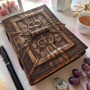 May include: A brown leather journal with an embossed design featuring a central bow and geometric patterns. The journal is closed, with a visible clasp. A black pen lies nearby, suggesting its use for writing. The overall aesthetic is rustic and detailed.