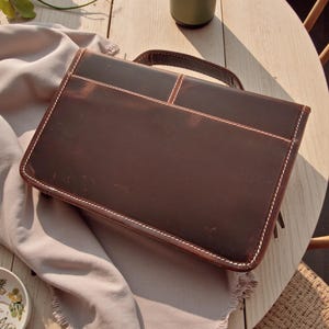 May include: A brown leather briefcase with a handle, white stitching, and a rectangular shape. The briefcase is sitting on a light-colored surface. The briefcase has a handle and a pocket on the front.