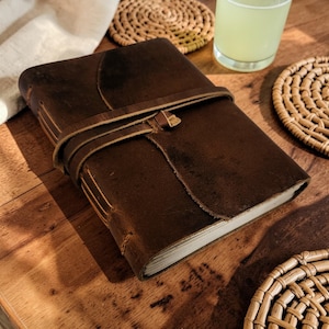 May include: A brown leather journal with a wrap-around strap closure. The journal has a textured cover and visible stitching along the spine. The strap is secured with a small loop. The journal is on a wooden surface.