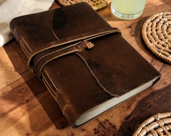 May include: A brown leather journal with a wrap-around strap closure. The journal has a textured cover and visible stitching along the spine. The strap is secured with a small loop. The journal is on a wooden surface.