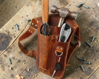 May include: A brown leather tool pouch featuring multiple pockets and loops. It contains a metal hammer, a pair of black-handled shears with a wooden handle, a utility knife, and a metal key ring with a black key attached to a D-ring.